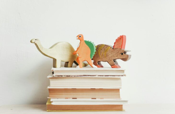 Colorful wooden dinosaurs on top of a stack of hardcover books on a light background.